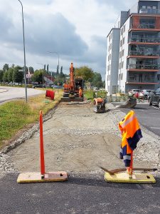 grusväg med grävmaskin och arbetare i orange klädsel som arbetar. Höghus till höger i bild.