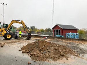 Grävmaskin, gruhög, rött hus på återvinningsstation i bakgrunden.