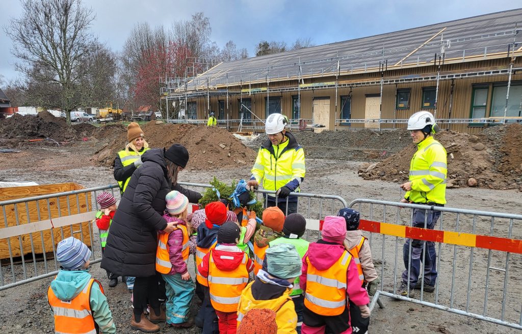 Barn i skyddsväst och färgglada kläder bakom staket på byggplats. Hus i bakgrunden och byggarbetare vid staketet.