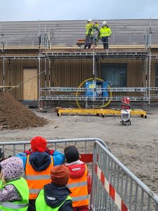 Barn och byggarbetare på byggplats. Grusplan och hus i bakgrunden.