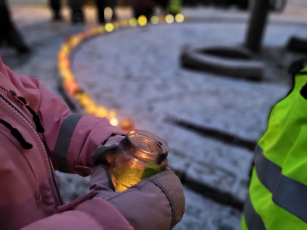 Barnhand med vante håller ljuslykta. Snö och fler ljuslyktor i bakgrunden.